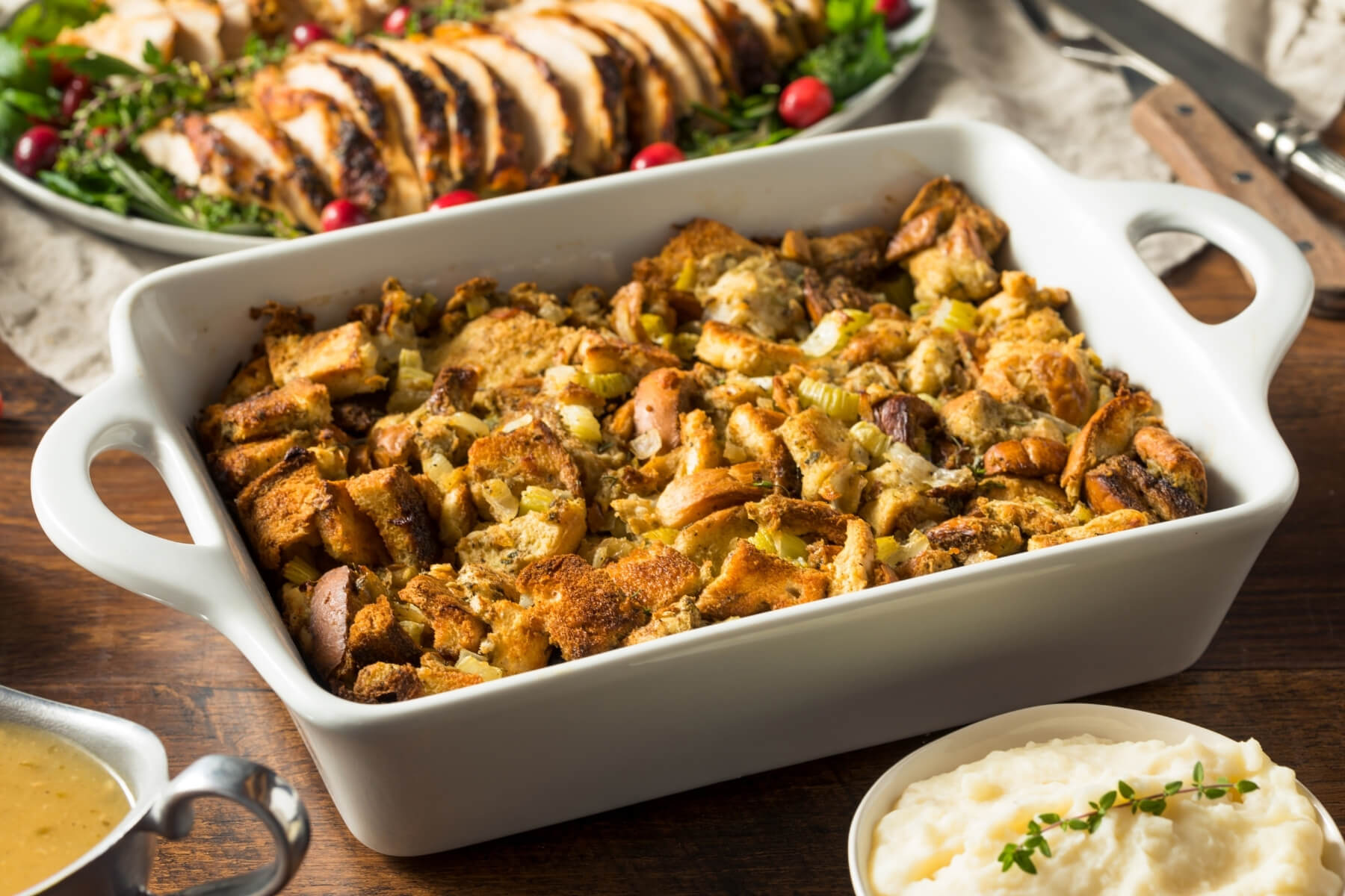 Baking dish of herb stuffing next to sliced turkey and mashed potatoes